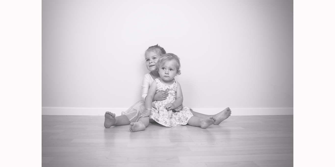 Familienshooting in Kerpen Zwei Kinder sitzen auf dem Boden, eines umarmt das andere, in einem hellen Raum.