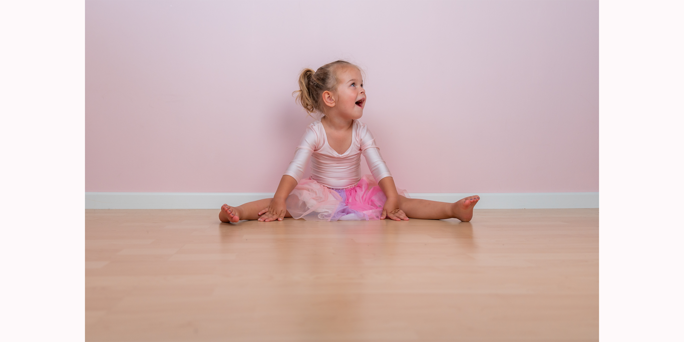 Ein kleines Mädchen sitzt mit gespreizten Beinen auf dem Boden vor einer rosa Wand.