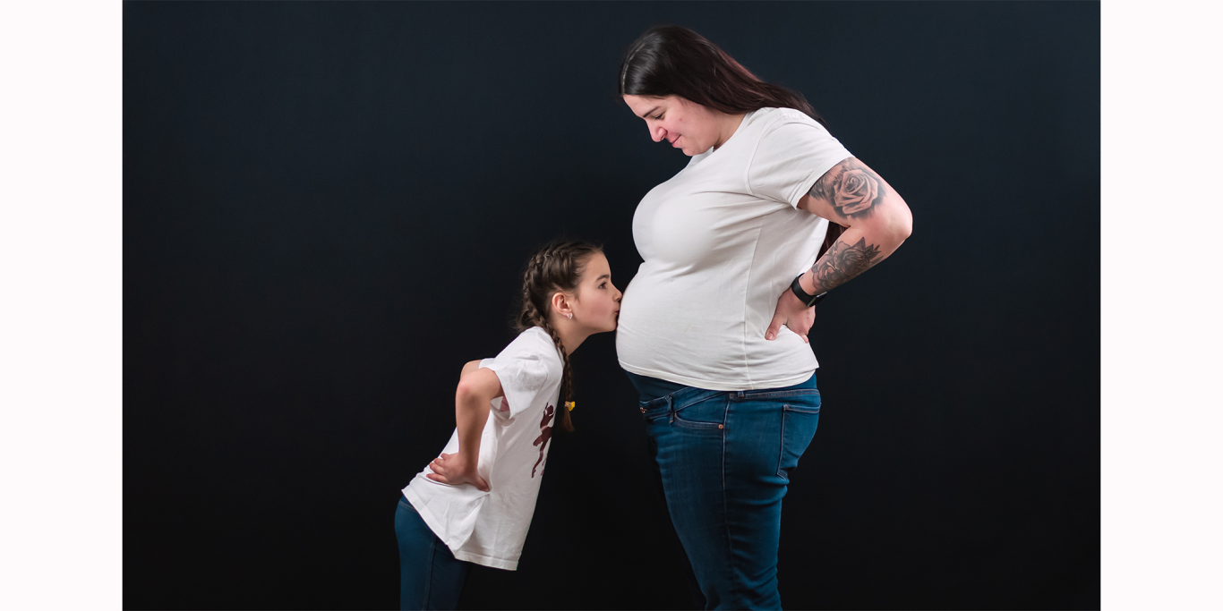 Babybauchshooting in Kerpen Eine schwangere Frau steht neben einem kleinen Mädchen, das neugierig auf ihren Bauch schaut.