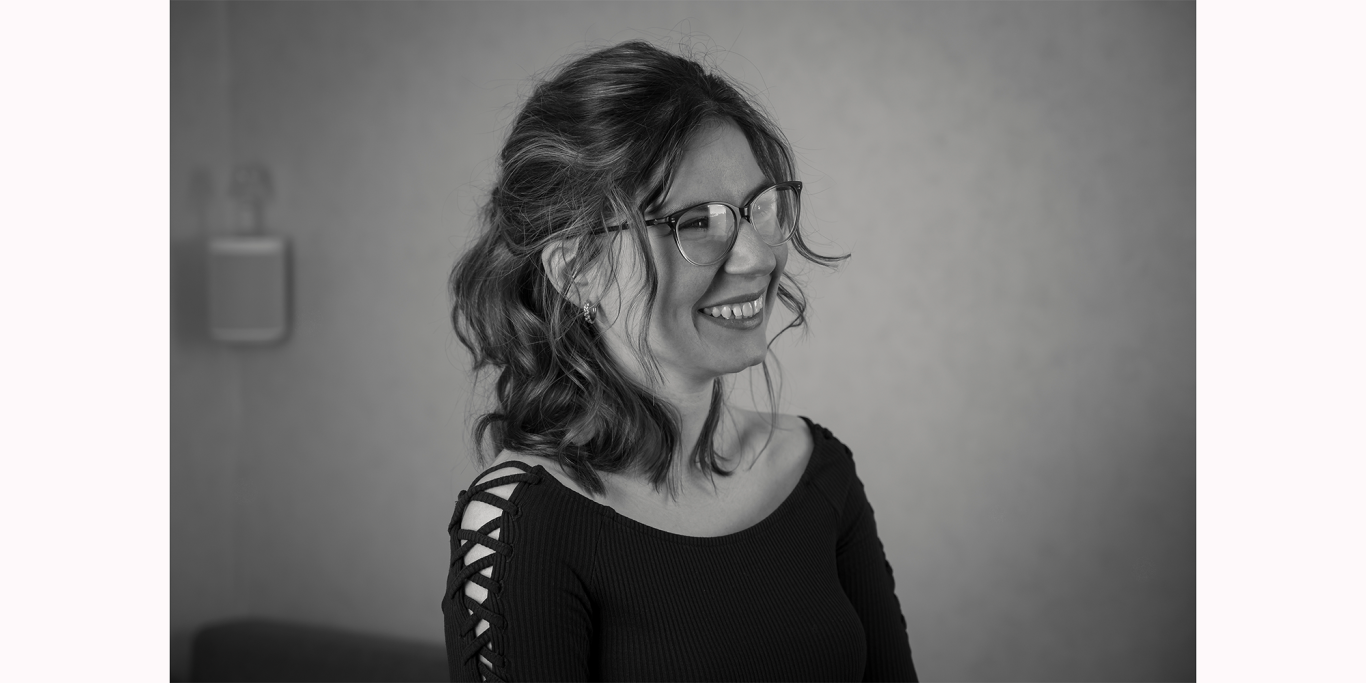 Lächelnde Frau mit lockigem Haar und Brille, in einem Raum mit neutralem Hintergrund.