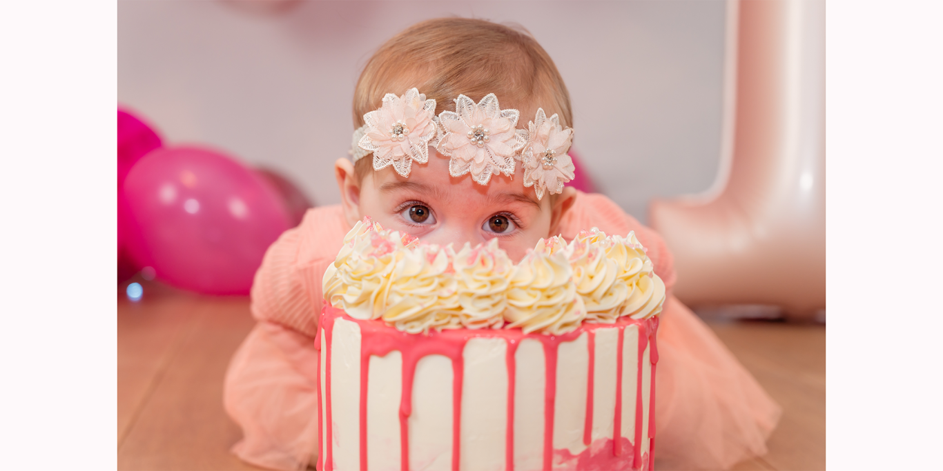 Ein Baby mit Blumenkranz sitzt vor einer Torte mit pinker Glasur und Sahne.
