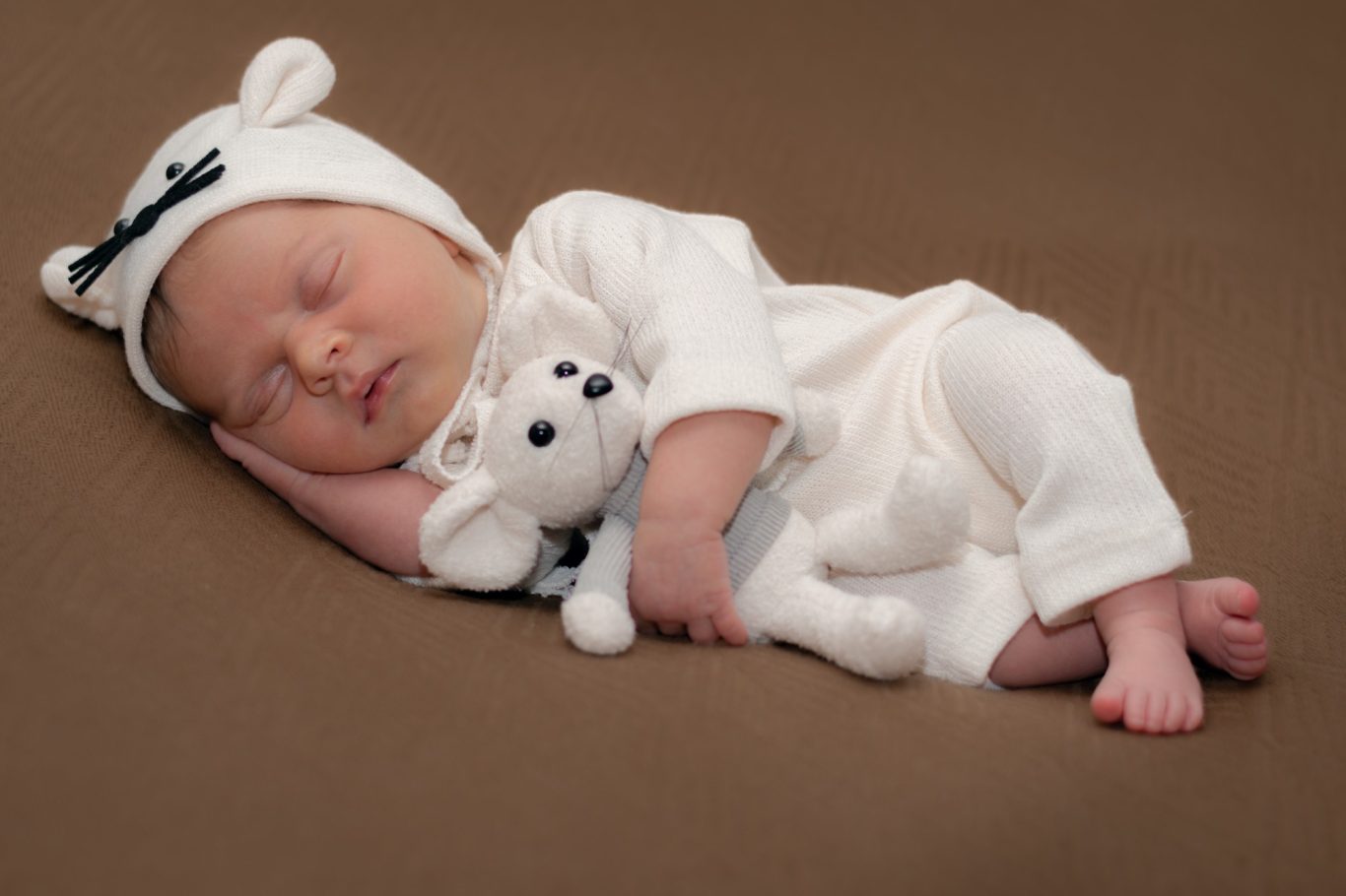 Neugeborenes Baby in einem weißen Outfit, schläft mit einem Stofftier in den Armen.