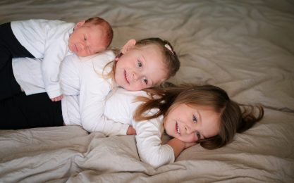 Drei Kinder liegen auf einem Bett, eines auf dem Rücken des anderen, fröhlich lächelnd.